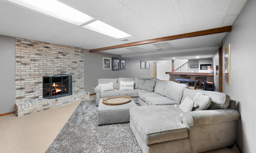 Modern basement renovation featuring a plush sectional sofa, classic brick fireplace, and custom bar space.